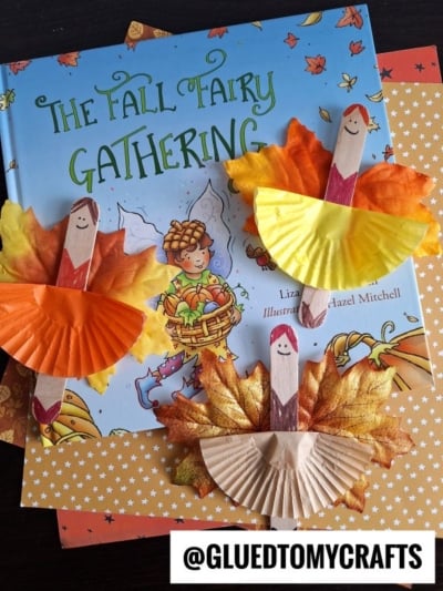 Three handmade fall crafts using popsicle sticks, paper cupcake liners, and artificial leaves arranged on top of the autumn-themed children’s book “The Fall Fairy Gathering.” Each smiling turkey resembles a playful leaf fairy ballerina.