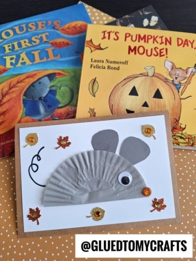 A cupcake liner mouse—handmade with a gray cupcake liner, googly eye, and paper ears—sits on a table surrounded by two autumn-themed children’s books, Mouse’s First Fall and It’s Pumpkin Day, Mouse!.