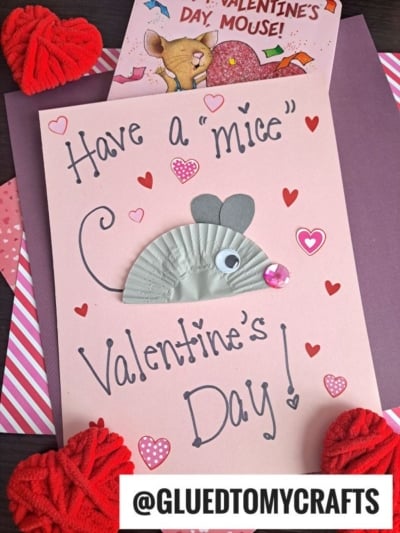 A handmade Valentine’s card features a cupcake liner mouse crafted from a gray liner, googly eye, and pink sequin on pink paper, with “Have a ‘mice’ Valentine’s Day!” surrounded by heart stickers. @GLUEDTOMYCRAFTS is at the bottom.