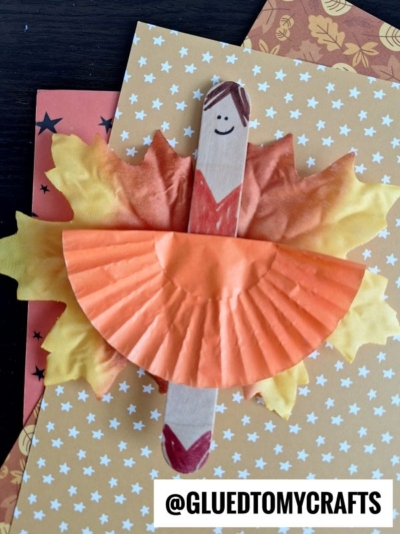 A fall-themed leaf fairy ballerina craft made from a wooden stick, artificial leaves, and an orange cupcake liner dress sits on patterned paper. The stick has a drawn face and hair, and the craft is tagged @GLUEDTOMYCRAFTS.