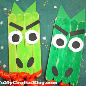 Two green popsicle stick dragon faces made from sticks, paper, and curled red ribbon for fire, displayed on a green background with white star decorations.