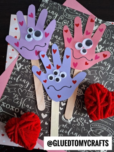 Three Monster Handprint Puppets made from colored paper, googly eyes, and heart stickers are attached to craft sticks. Displayed on Valentine-themed backgrounds, these fun crafts are perfect for celebrating your mommy's little monster.
