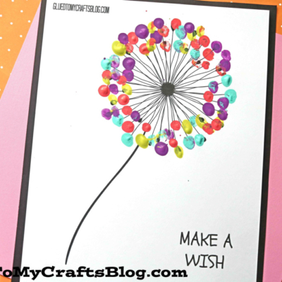 A white sheet with a printed Thumbprint Dandelion features colorful thumbprints as seeds. Paint pots and a brush sit beside it. The text reads MAKE A WISH