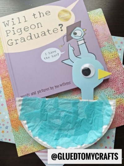 A children’s book titled “Will the Pigeon Graduate?” is next to a blue paper plate pigeon craft with construction paper, googly eye, and beak, on a colorful patterned background. @GLUEDTOMYCRAFTS appears at the bottom.