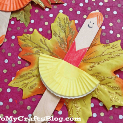 A fall-themed craft featuring a leaf fairy ballerina made from a popsicle stick, cupcake liner dress, and artificial leaves, set on a pink polka dot background. The figure has a drawn-on smiling face and brown hair.