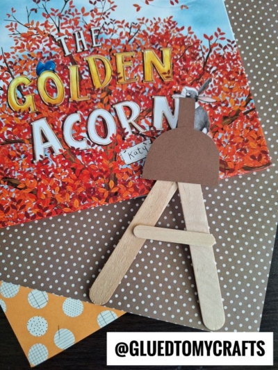 A popsicle stick acorn craft made from brown paper sits on patterned paper, with the book The Golden Acorn in the background. A label reads @GLUEDTOMYCRAFTS at the bottom right.