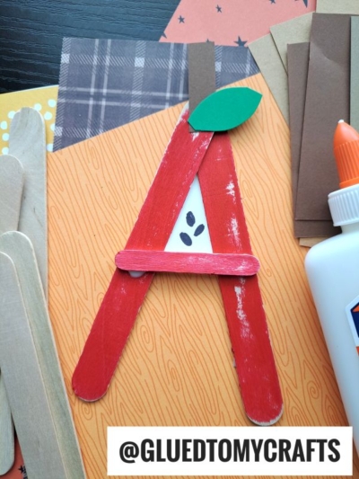 A popsicle stick "A" craft project, shaped from red sticks with a green paper leaf and brown stem to resemble an apple, sits on patterned paper. Glue and extra craft sticks are nearby for this fun alphabet-themed activity.