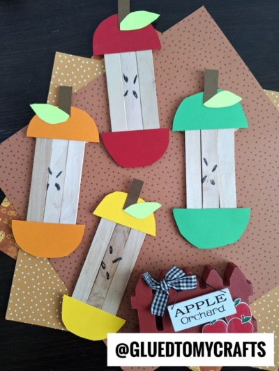 Four craft stick apple core crafts made from popsicle sticks and colorful paper are displayed on patterned paper. Each apple has a different color—red, green, orange, and yellow—with paper leaves and seeds drawn in the center.