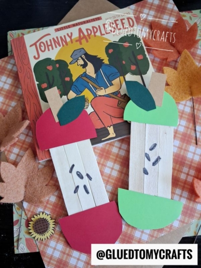 Two popsicle stick apple core crafts made from colored paper and craft sticks sit on fall-themed paper. In the background is the “Johnny Appleseed” book, felt leaves, and a small sunflower decoration. @GLUEDTOMYCRAFTS watermark is visible.