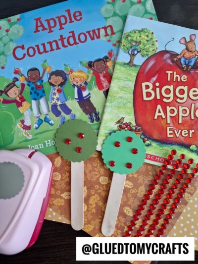 Two children’s books, Apple Countdown and The Biggest Apple Ever, are surrounded by apple tree craft for kids supplies, including popsicle stick apple trees, red gem stickers, patterned paper, and a pink paper punch. @GLUEDTOMYCRAFTS text label at bottom.