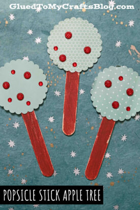 Three craft stick apple trees with red sticks and round green tops dotted with red glitter, arranged on a blue textured background—a fun apple tree craft for kids. Text: POPSICLE STICK APPLE TREE.
