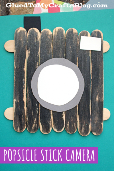 A popsicle stick camera crafted from black-painted sticks features a white and gray paper lens and small black and white button details, displayed on a teal background. Text reads POPSICLE STICK CAMERA.