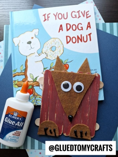 A childrens book titled If You Give a Dog a Donut sits with a bottle of Elmers glue, blue and patterned paper, and a popsicle stick dog craft made from painted sticks and brown felt. @GLUEDTOMYCRAFTS is written at the bottom.