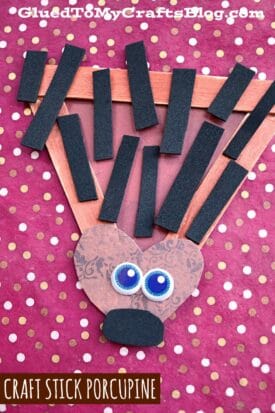 A popsicle stick porcupine craft made from a triangular frame, with a heart-shaped paper face, googly eyes, black nose, and black foam rectangle quills on a red polka-dot background. Text reads CRAFT STICK PORCUPINE.