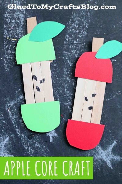 Two apple core crafts made from craft sticks and colored paper—a charming craft stick apple core—one with green paper tops and bottoms and the other with red, both featuring green leaves and drawn seeds, displayed on a dark background.