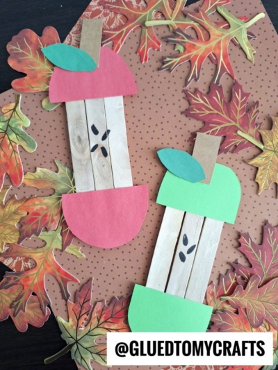 Two popsicle stick apple core crafts made from green and red paper with marker seeds are displayed on brown polka dot paper, surrounded by decorative fall leaves.