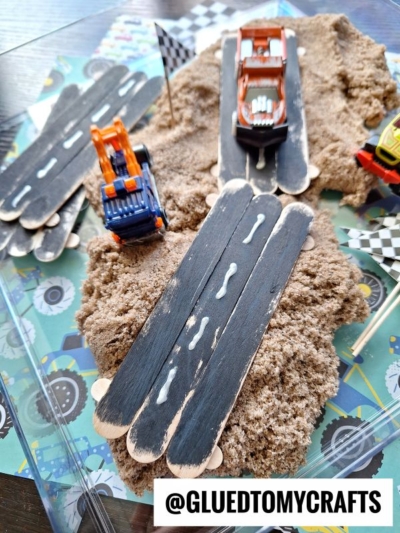 Toy cars zoom along a popsicle stick race track, painted black and set over kinetic sand. Checkered flags add flair, while a bold sign reads @GLUEDTOMYCRAFTS, creating the perfect miniature racing scene.