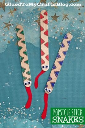Three craft stick snakes decorated with zigzag patterns in green, red, and blue, each with googly eyes and a red paper tongue, displayed on a blue starry background. Text reads POPSICLE STICK SNAKES.
