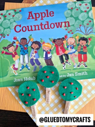 A children's book titled Apple Countdown is shown with three Clothespin Felt Apple Trees made from felt and popsicle sticks placed in front. The book cover features diverse children holding apples.