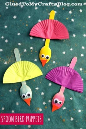 Three colorful cupcake liner bird puppets made from painted wooden spoons, googly eyes, and orange paper beaks are displayed on a star-patterned background. Text reads, Spoon Bird Puppets.