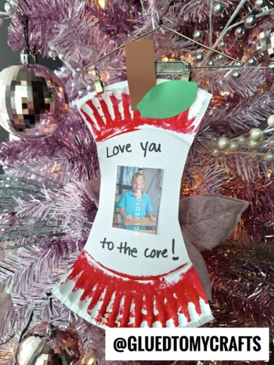 A handmade paper plate apple core photo frame ornament with red edges, a green leaf, and the message Love you to the core! hangs on a pink Christmas tree. A photo is displayed in the center. The handle @GLUEDTOMYCRAFTS is shown below.