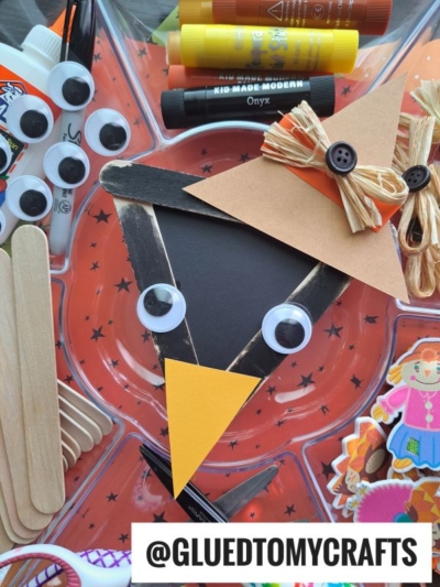 A craft tray holds googly eyes, glue sticks, craft sticks, and a DIY scarecrow face made from paper, a googly eye, a button hat, and straw. Beside it sits a Popsicle Stick Black Crow ready to join the fun fall crafting. @GLUEDTOMYCRAFTS text label is at the bottom.