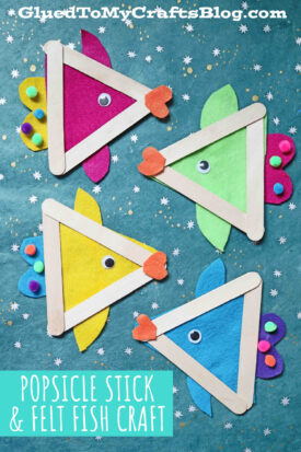 Four colorful popsicle stick fish crafts made from triangle-shaped sticks and felt, decorated with googly eyes, felt fins, and pom-pom noses, are arranged on a blue star-patterned background.