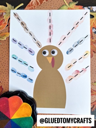 A turkey craft made from brown paper with googly eyes, a red wattle, and colorful thumbprint turkey feathers sits on a white sheet. Nearby are a flower-shaped paint palette and autumn leaves. @GLUEDTOMYCRAFTS is written at the bottom.