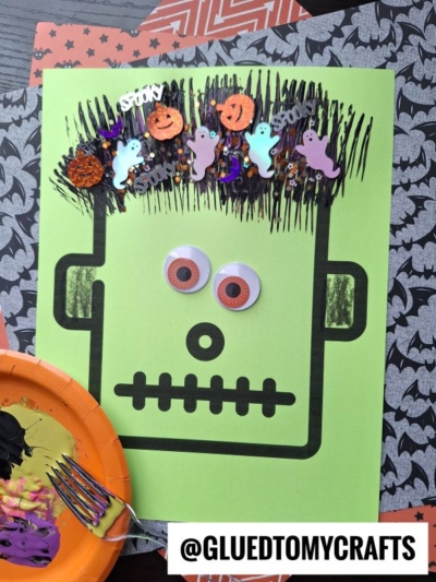 A green paper craft of Frankenstein’s monster face with fork painted monster hair, googly eyes, black marker features, and colorful Halloween confetti. Crafting materials and a painted plate are nearby. @GLUEDTOMYCRAFTS is written at the bottom.
