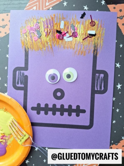 A purple paper craft monster face with googly eyes, fork painted monster hair in yellow, and Halloween confetti on a star-patterned background. @GLUEDTOMYCRAFTS text label appears at the bottom right.