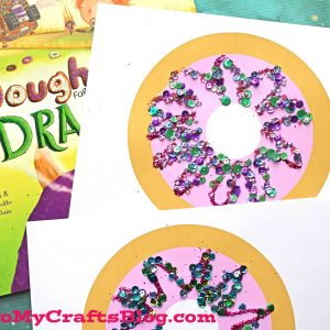 Two handmade paper doughnut crafts decorated with colorful sequins and glitter sit on white paper, with the book Doughnuts for Dragons partially visible underneath. The website GluedToMyCraftsBlog.com appears at the bottom. A fun doughnut kid craft!.