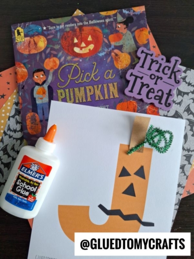 A Halloween-themed craft scene with an Elmer’s glue bottle, a Pick a Pumpkin book, a Trick or Treat sign, and a "j is for jack-o-lantern" paper pumpkin craft on white paper. The Instagram handle @GLUEDTOMYCRAFTS is at the bottom.