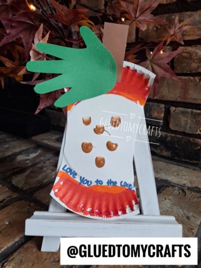 Paper plate apple core craft featuring a green handprint leaf, a brown stem, and drawn seeds; displayed on a small white easel. Text at the bottom reads @GLUEDTOMYCRAFTS.