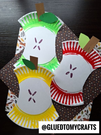 Three paper plate apple core crafts in red, green, and yellow are displayed on patterned papers. Each features a brown paper stem, green leaf, and drawn seeds. The text @GLUEDTOMYCRAFTS appears at the bottom.