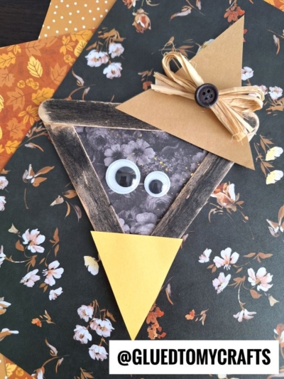 A handmade witch craft made from a wooden triangle frame, yellow paper hat with a button and raffia bow, plastic googly eyes, and yellow paper nose—plus a Popsicle Stick Black Crow—on floral and autumn-patterned paper. @GLUEDTOMYCRAFTS at bottom.