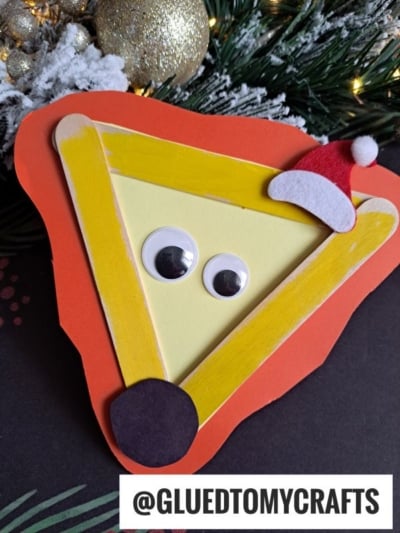 A handmade Christmas card featuring a triangular reindeer face crafted in the style of a craft stick lion, with googly eyes, a red nose, and a tiny Santa hat, set against festive greenery. The @GLUEDTOMYCRAFTS handle appears at the bottom.