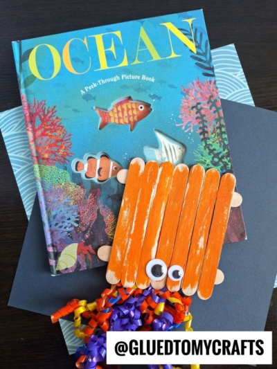 A children’s book titled OCEAN sits on a table, topped with an adorable popsicle stick octopus craft made from googly eyes and curled ribbon. A fish cutout and the @GLUEDTOMYCRAFTS handle are also visible.