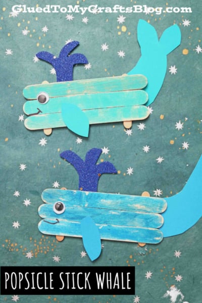 Two craft stick whale creations made from painted popsicle sticks, blue paper tails and fins, googly eyes, and sparkly blue water spouts are displayed on a green background with star and splatter designs. Text reads “POPSICLE STICK WHALE.”.