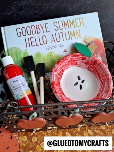 A basket with the book Goodbye Summer, Hello Autumn, a bottle of red acrylic paint, two foam brushes, a paper bowl apple craft, and autumn-themed decorations. @gluedtomycrafts is displayed at the bottom.