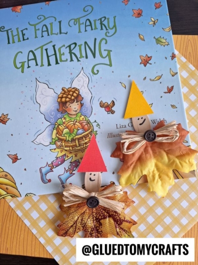 A children’s book titled The Fall Fairy Gathering is displayed with two scoop stick leaf elves crafts, made from artificial leaves, buttons, raffia bows, and paper hats, both with smiling faces. The username @GLUEDTOMYCRAFTS is overlaid.