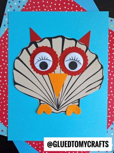 A seashell owl made from a seashell-shaped cutout, red and orange paper for eyes, beak, and ears, glued onto a blue background with a red star-patterned paper behind it. @GLUEDTOMYCRAFTS is printed at the bottom.