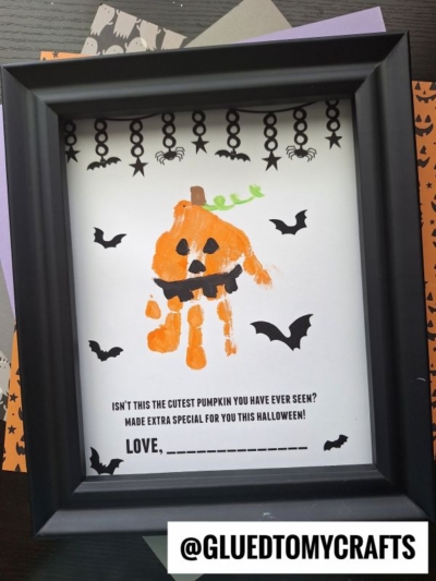 A framed Halloween craft features a Handprint Jack-O-Lantern made from an orange-painted handprint, complete with black facial features and a green stem. Flying bats and spider decorations surround it, with space below for a personalized signature.