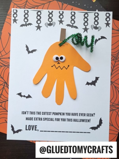 Handprint Jack-O-Lantern Halloween craft featuring a googly eye, pipe cleaner stem, and zigzag mouth on paper with festive Halloween-themed decorations and a special message for kids. This handprint pumpkin keepsake is perfect for little ones to create lasting memories. @GLUEDTOMYCRAFTS at the bottom.