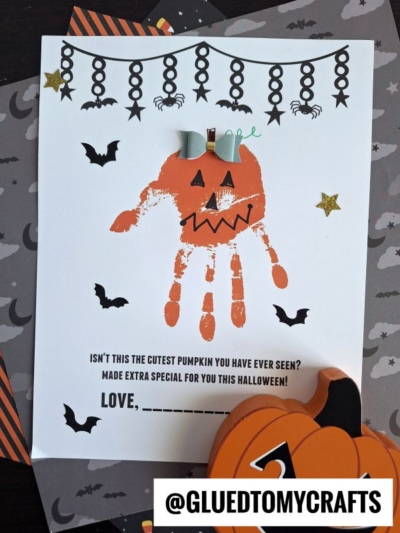 A Halloween craft featuring a Handprint Jack-O-Lantern with a jack-o’-lantern face, paper bow, and bat, star, and spider decorations. The text reads, “Isn’t this the cutest handprint pumpkin keepsake you have ever seen?”.