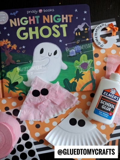 Childrens book Night Night Ghost with pink and white ghost crafts, including Paper Plate Ghosts made from napkins and cupcake liners, black dot eyes, orange polka-dot paper, Elmers glue bottle, and crafting supplies on a table.