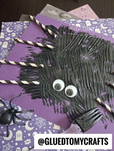 Purple paper with black paint spread in a spiky shape, google eyes, paper straw spider legs made from striped straws, a small plastic spider, and patterned Halloween paper underneath. This playful paper straw spider craft has GLUEDTOMYCRAFTS written at the bottom.