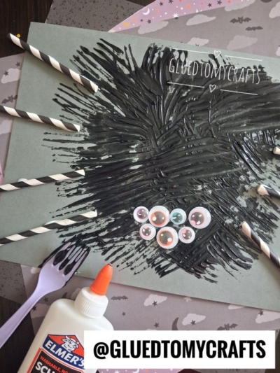 Black paint spread on gray paper, with googly eyes and striped straws attached to look like paper straw spider legs, creating a fun straw spider craft. Craft glue and @GLUEDTOMYCRAFTS label are also visible. Bat-patterned background paper is underneath.