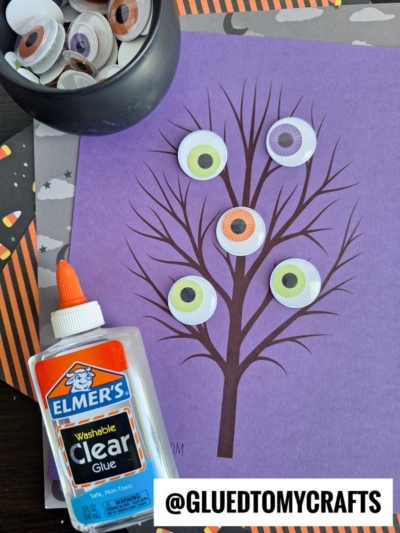 A purple paper with a brown wiggle eye tree design has colorful googly eyes glued on its branches. A bottle of clear glue and a bowl of googly eyes are nearby. Halloween-themed paper is in the background.
