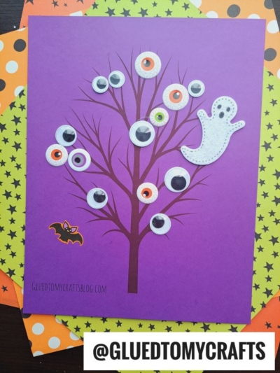Purple paper with a brown wiggle eye tree, decorated with googly eyes, a white paper ghost, and a bat sticker. The background has polka dot and star designs. A label reads @GLUEDTOMYCRAFTS.