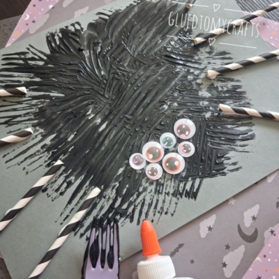 A gray paper with black paint spread like a spider, googly eyes, and paper straw spider legs attached with glue, plus a plastic fork. The background features decorative papers. @GLUEDTOMYCRAFTS is written at the bottom.